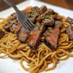 "Plate of sliced garlic butter steak served on top of glossy stir-fried noodles with scallions and garlic